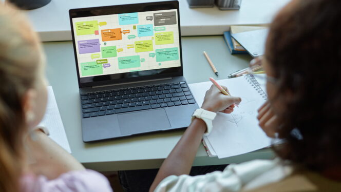 Over shoulder view of two students using computer together in school classroom with colorful charts on screen