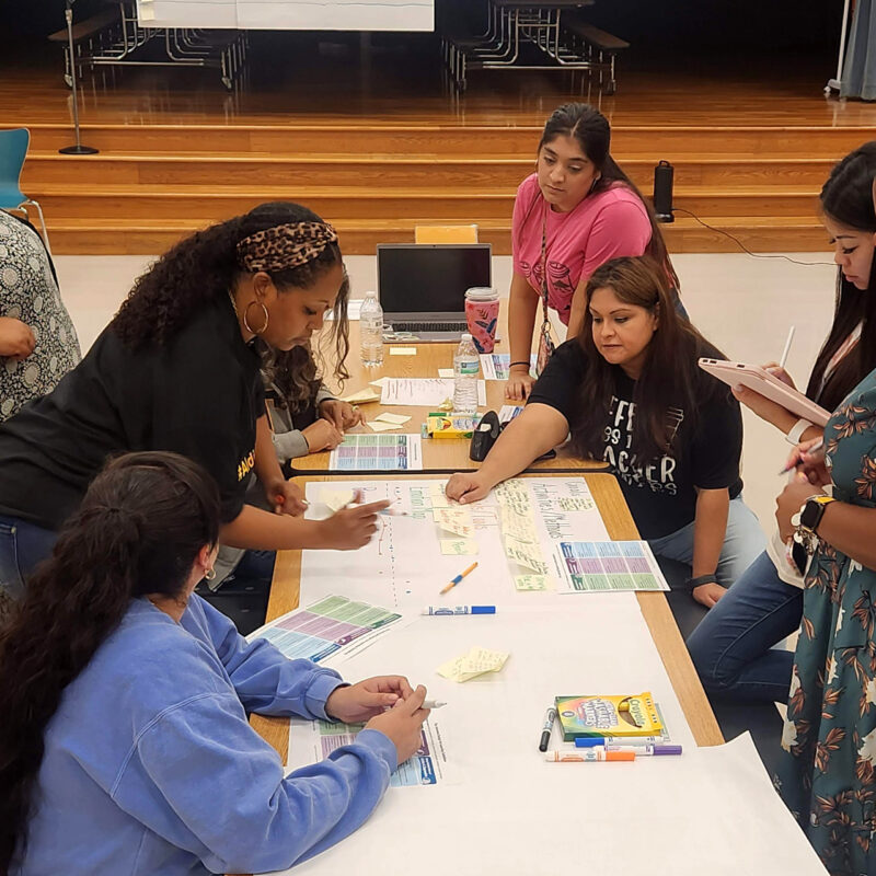 Team of educators working together at a long table covered with markers, guidelines, and paper.
