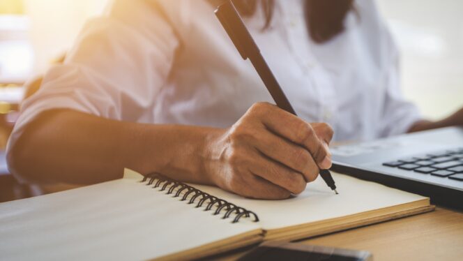 A person writing in a notebook at a desk with a laptop and phone