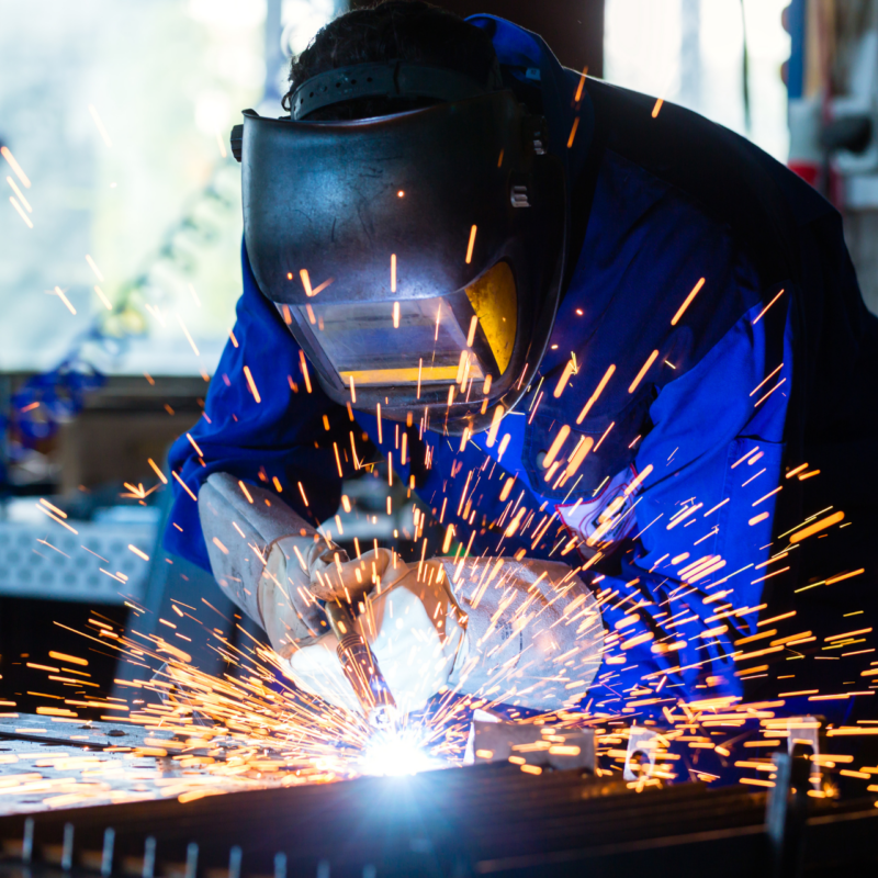 Photo of a welder in a blue jumpsuit and mask welding, with sparks flying.