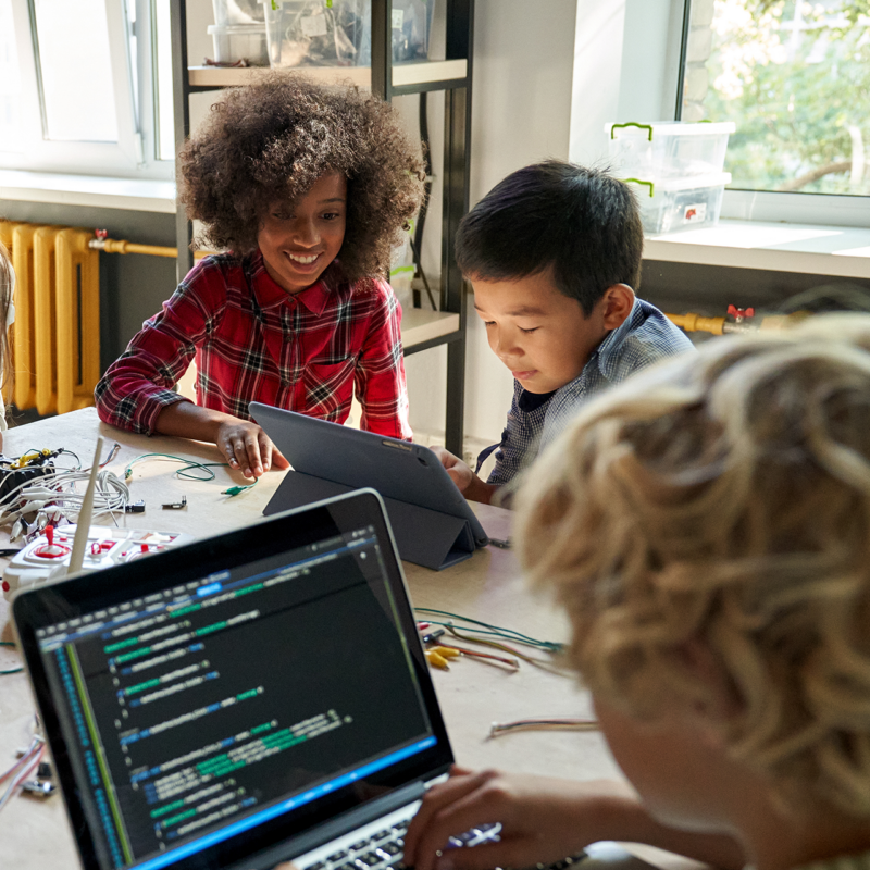 Three engaged students working on an robotics project in a classroom, with programming code visible on a laptop, and electronics components on the table.
